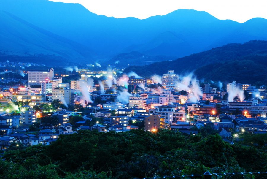 Panoramic view of Beppu city at dusk, showing numerous plumes of hot spring steam rising above the illuminated buildings against a deep blue mountain backdrop.