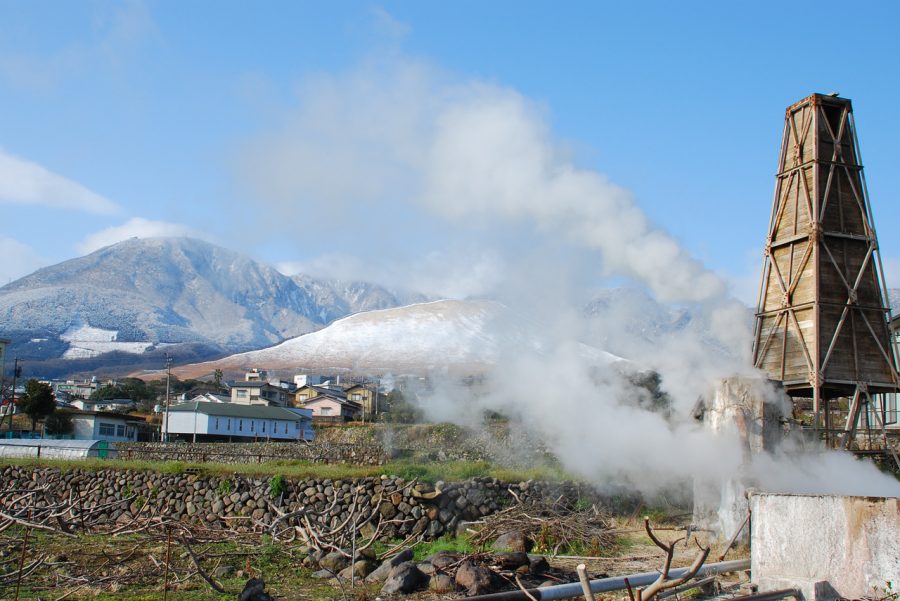 Geothermal steam vent in Beppu, Japan, with a tall wooden structure emitting hot steam against a backdrop of snow-dusted mountains.