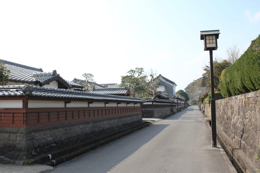 A quiet street in Obi castle town, lined by white walls with tiled roofs and stone foundations, and a high green hedge on the right.