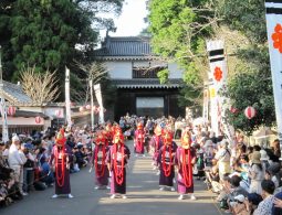 Obi Castle Festival parade with participants in traditional purple costumes and fox masks marching through a crowded street toward a castle gate.