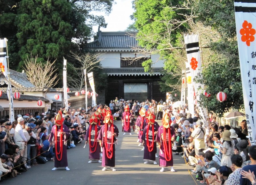 Obi Castle Festival parade with participants in traditional purple costumes and fox masks marching through a crowded street toward a castle gate.