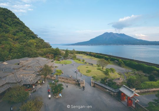 An elevated view of Sengan-en Garden and buildings, with the open water of Kagoshima Bay and the Sakurajima volcano in the distance at sunset.