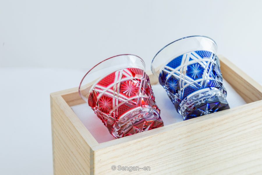 Two richly cut Kiriko glass tumblers, one red and one blue, likely representing Satsuma Kiriko, displayed in a wooden box.