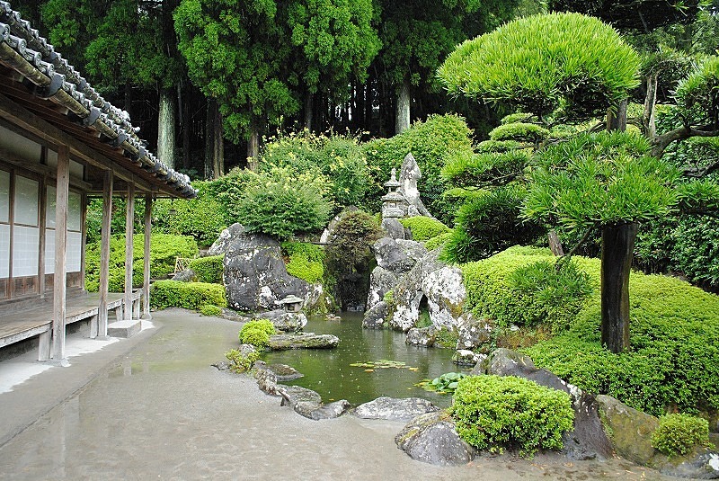 A traditional Japanese dry landscape garden (karesansui or pond style), featuring a small pond, sculpted pine trees, rocks, and a veranda.