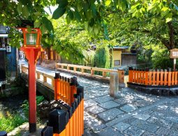 Small stone bridge over Shirakawa River in Kyoto on a sunny day, framed by lush green trees and traditional orange railings.