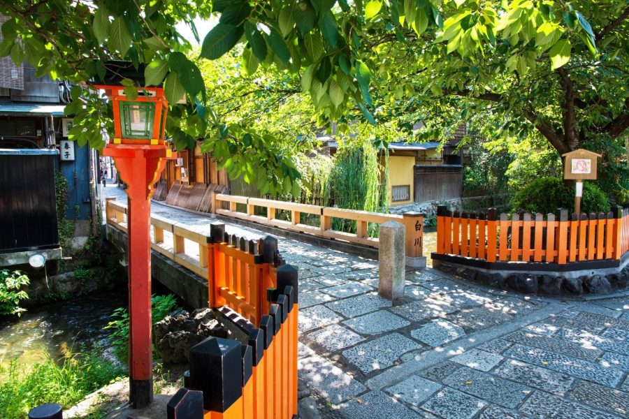 Small stone bridge over Shirakawa River in Kyoto on a sunny day, framed by lush green trees and traditional orange railings.