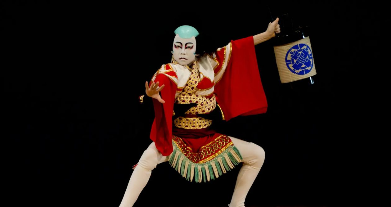 A Kabuki performer in dramatic red, white, and gold costume and kumadori makeup, holding a lantern against a black background.