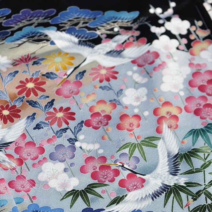 Close-up of a colorful Japanese kimono fabric featuring a vibrant pattern of white cranes, plum blossoms, chrysanthemums, bamboo, and pine needles on a light blue and black background.