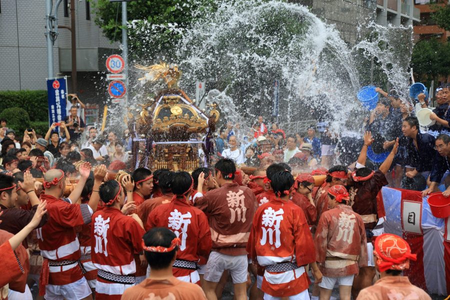 Scene from the Fukagawa Hachiman Festival, widely known as the "Water-throwing Festival." Bearers wearing matching red happi coats carry a portable shrine (mikoshi) while being vigorously splashed with water from buckets by the crowd.