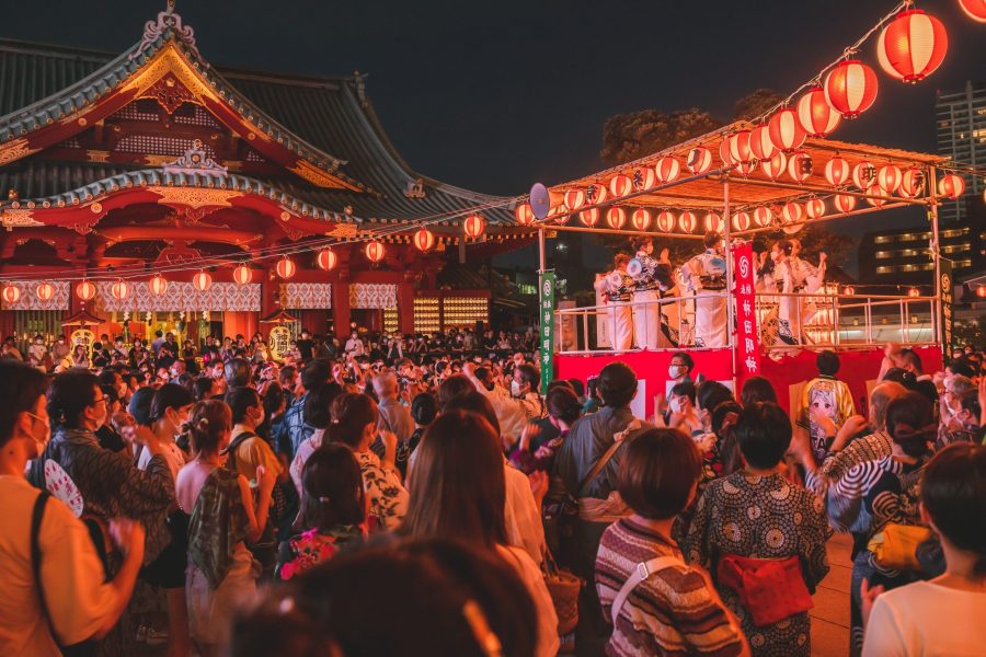 Night scene at Kanda Myoujin Shrine. A lively Bon Odori (dance festival) is taking place in the shrine precincts at night. A large crowd, many wearing yukata, dances in a circle around a raised wooden stage (yagura) illuminated by rows of glowing red and white lanterns. The majestic main hall of the shrine stands in the background, beautifully lit up against the night sky.