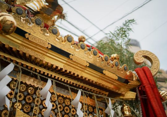 Close-up of the intricate gold decorations on a portable shrine (mikoshi). Detailed metalwork featuring crests and wave patterns adorns the black lacquered roof, highlighting the exquisite craftsmanship.
