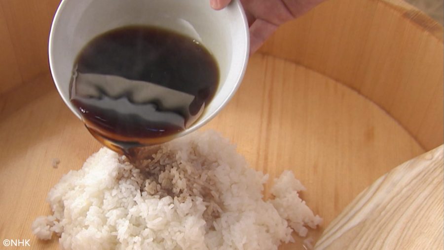 Mixing red vinegar (Akazu) into sushi rice. A dark, amber-colored vinegar is being poured from a white bowl onto steaming white rice in a wooden tub (hangiri).
