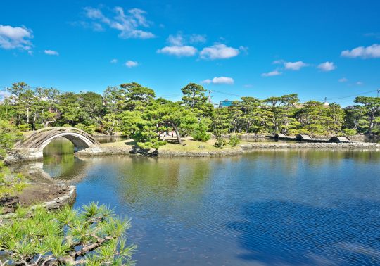 View of a traditional Japanese garden featuring a pond, pine trees, and a stone arched bridge under a blue sky.