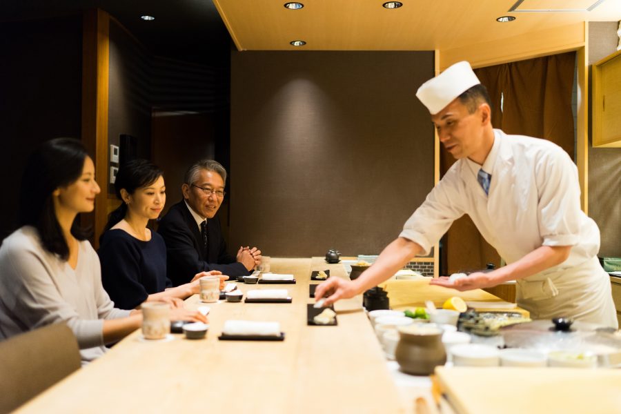 A sushi chef in a white uniform serving sushi to customers seated at a wooden counter in a high-end restaurant.