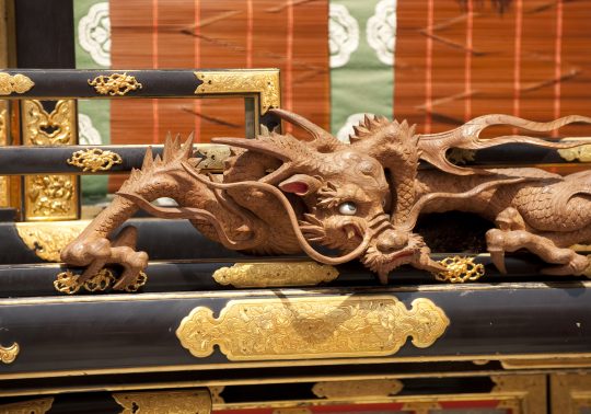 Close-up of a wooden dragon carving on a portable shrine (mikoshi). The unpainted wood sculpture features intricate details in the scales and claws, with an expressive eye that seems to glare. It is set against black lacquered wood adorned with gold fittings.