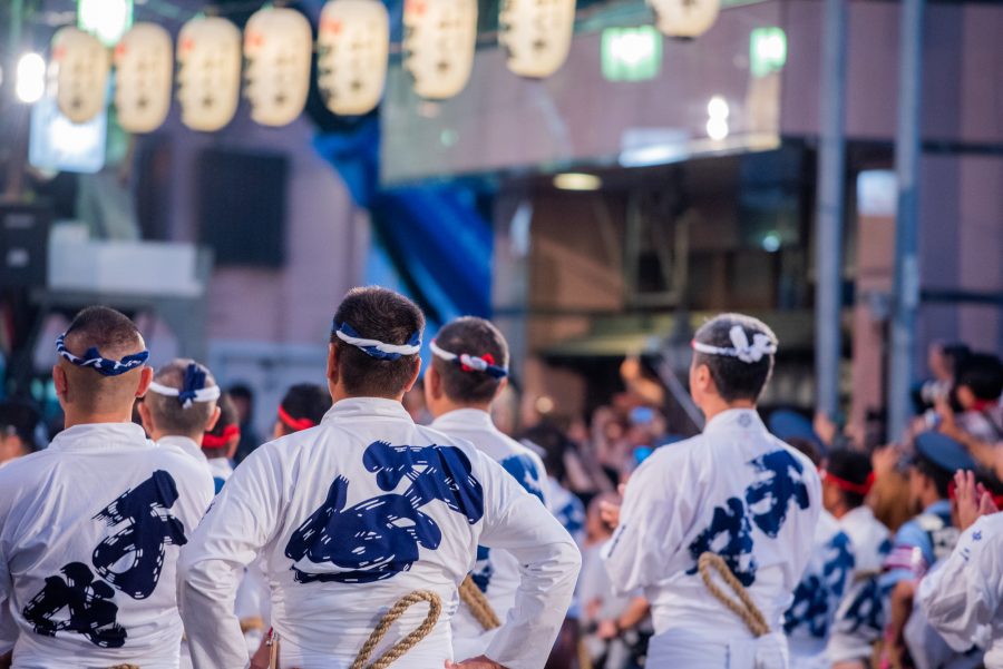 Scene from the Hakata Gion Yamakasa Festival in Fukuoka. A rear view of men wearing traditional white happi coats with blue designs, standing in a row under the glow of paper lanterns.