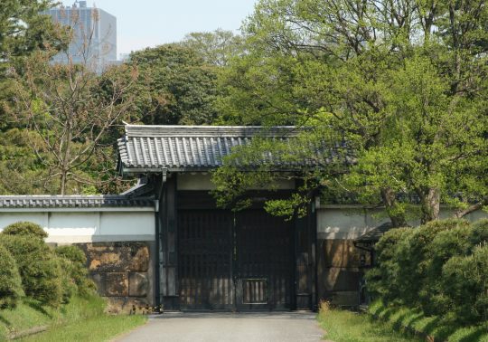 Hanzomon Gate of the Imperial Palace (formerly Edo Castle). A strictly guarded wooden gate set into massive stone walls, surrounded by deep moats and lush greenery.