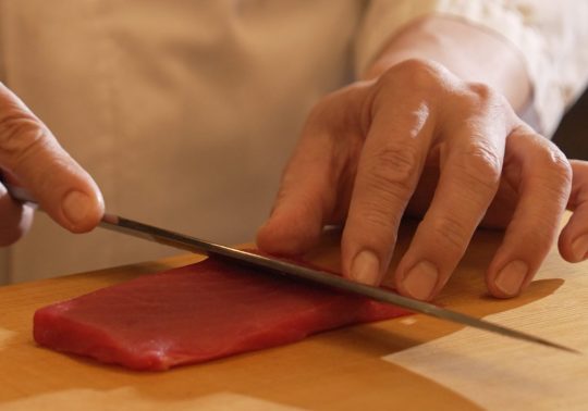 Close-up of a sushi chef slicing a block of red tuna (maguro) with a long, sharp sashimi knife on a wooden cutting board.