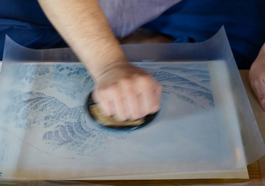 Close-up of a craftsman's hands using a baren tool to press a woodblock print of The Great Wave.