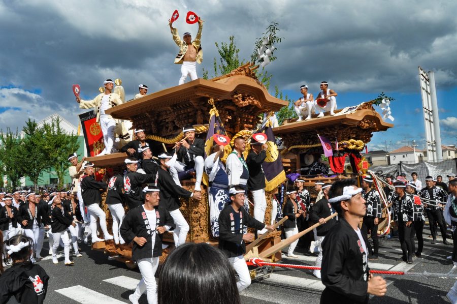 Scene from the energetic Kishiwada Danjiri Festival in Osaka. A massive wooden float (danjiri) with intricate carvings is being pulled by a large crowd. Men stand on the roof, with the "daikugata" (carpenter) dancing with fans to direct the float's movement.
