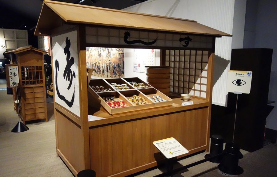 Replica of an Edo-period sushi stall displayed in a museum. The wooden food stand features a noren curtain with the character for "Sushi" and displays wooden boxes filled with various types of nigiri sushi.