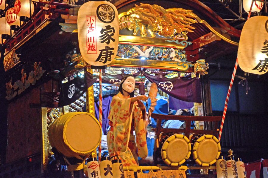 m the Kawagoe Festival. A magnificent festival float (dashi) glows under the light of paper lanterns at night. A dancer wearing a traditional mask (Okame) and kimono performs to the rhythm of drums (taiko) and flutes (ohayashi) on the stage of the float. The lantern prominently displays the name "Ieyasu" (Tokugawa Ieyasu) and the crest of the Tokugawa family, symbolizing the deep connection to the Edo Shogunate.