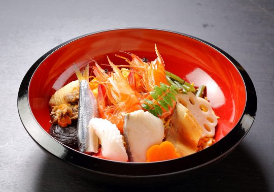 Bara-zushi (Scattered Sushi) served in a lacquer bowl. The sushi rice is topped with a colorful assortment of ingredients, including whole boiled shrimp, grilled fish (Spanish mackerel), lotus root, bamboo shoots, shiitake mushrooms, and thin strips of egg omelet.