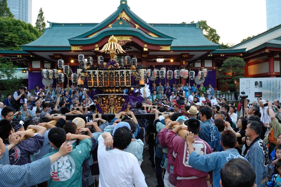 Modern-day scene of the Sanno Festival at Hie Shrine in Tokyo. A large crowd of bearers wearing traditional happi coats is energetically carrying an ornate portable shrine (mikoshi) in front of the main shrine building.