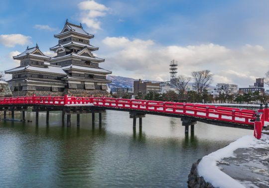 Matsumoto Castle in winter featuring a red bridge over the moat and snow.