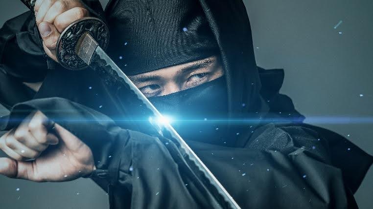 A close-up, dramatic photo of a person dressed in black ninja attire, holding a katana sword across the face, with only the eyes visible.